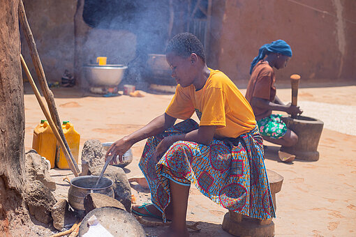 Eine ghanaische Teenagerin sitzt draußen auf einem Hocker und rührt in einem Topf, der auf einer Feuerstellt steht. Im Hintergrund bereitet eine ghanaische Frau in einem Tongefäß Essen zu