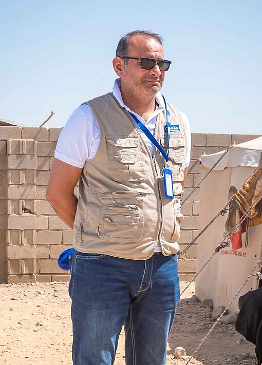 Ein Mann mit brauner Weste und Sonnenbrille steht auf einem sandigen Platz