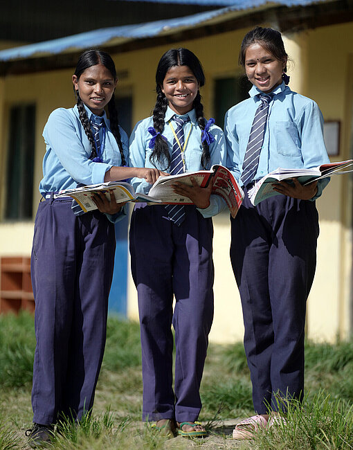 Drei Schülerinnen in Schuluniform aus Nepal lesen gemeinsam Bücher im Freien vor einem Schulgebäude
