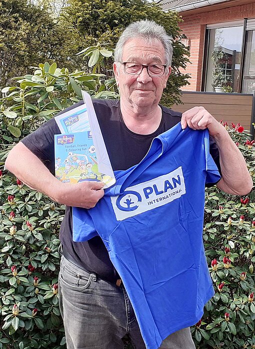 Ein Mann steht im Garten eines hauses und hält ein T-Shirt mit dem Plan-Logo hoch.