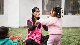 Eine Frau in einer rosa Weste spielt mit Kleinkindern auf einer Wiese. Ein Kind hält ihre Hände, während andere mit bunten Bällen spielen.