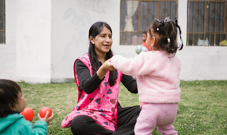 Eine Frau in einer rosa Weste spielt mit Kleinkindern auf einer Wiese. Ein Kind hält ihre Hände, während andere mit bunten Bällen spielen.