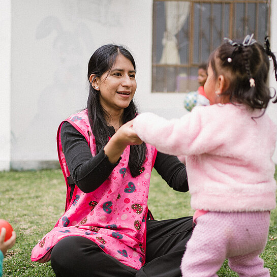 Eine Frau in einer rosa Weste spielt mit Kleinkindern auf einer Wiese. Ein Kind hält ihre Hände, während andere mit bunten Bällen spielen.