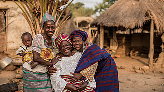 Eine glückliche Familie aus Togo posiert zusammen vor traditionellen Hütten. Eine ältere Frau sitzt in der Mitte, während ihre Tochter und Enkelkinder sie umarmen.