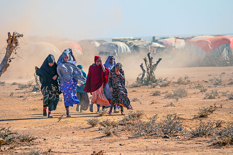 Eine Gruppe Mädchen in bunten Gewändern läuft durch eine staubige Wüstenlandschaft. Im Hintergrund ist eine Zelt-Siedlung zu erkennen.
