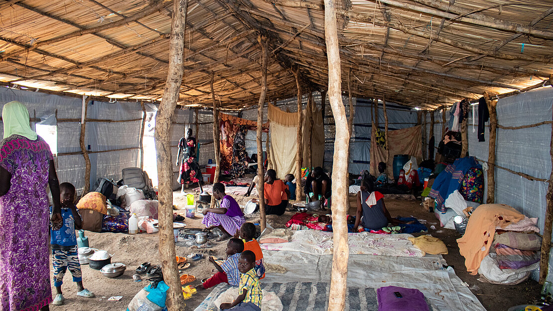 Im inneren eines großem Zeltes sitzen mehrere Menschen auf dem Boden auf Decken, eine Frau und ein Kind stehen links im Bild, an einigen Stöcken sind Tücher als Sichtschutz aufgehängt
