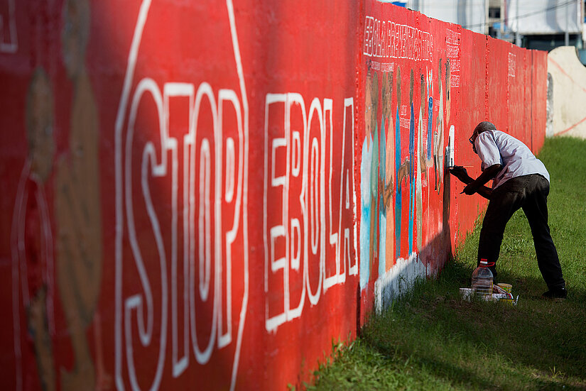 Ein Mann bemalt eine lange, rote Mauer mit großem weißen Schriftzug „STOP EBOLA“ und bunten Figuren. Die Mauer steht auf einer Wiese; im Hintergrund sind weitere Gebäude erkennbar.