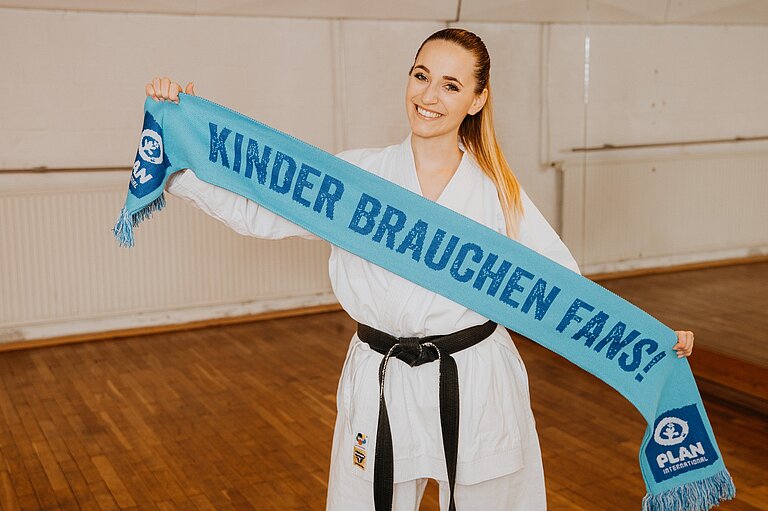 Eine Frau im Karateanzug hält lächelnd den „Kinder brauchen Fans“-Schal in die Kamera