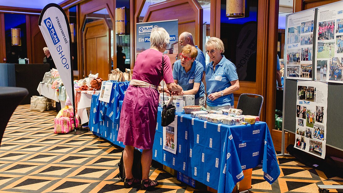 Drei Mitglieder der Plan-Aktionsgruppe Köln sitzen an einem Tisch und Tombola-Gewinnen und Informationen zu einem so unterstützten Projekt.
