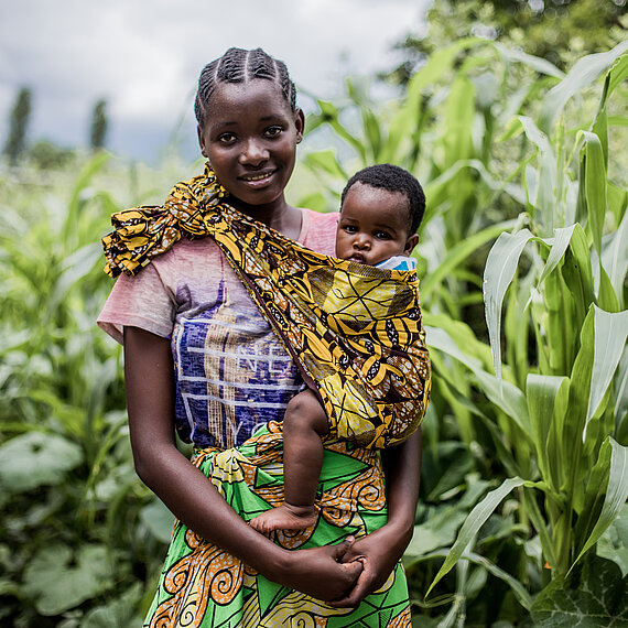 Junge Mutter mit ihrem Baby im Tragetuch steht lächelnd auf einem Feld mit hohem Grün in Sambia.