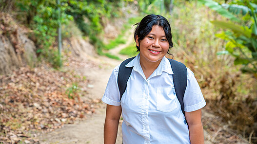 Junge Frau mit einem Rucksack auf dem Rücken ist im Grünen auf dem Weg zur Schule in El Salvador.