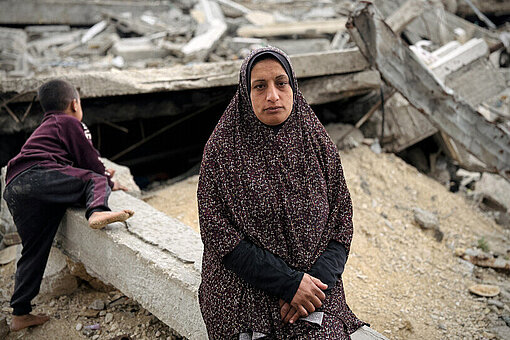 Eine Mutter aus Gaza steht vor den Trümmern ihres Hauses. Hinter ihr klettert ein kleiner Junge gerade auf ein Trümmerteil.