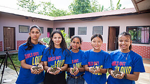 Fünf junge Frauen in Nepal tragen ‚Girls Out Loud‘-Shirts und halten Pflanzensetzlinge in den Händen