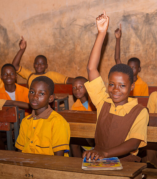Mehrere ghanaische Kinder sitzen in einem Klassenzimmer. Sie tragen Schuluniformen. Drei von ihnen, darunter ein Mädchen, melden sich