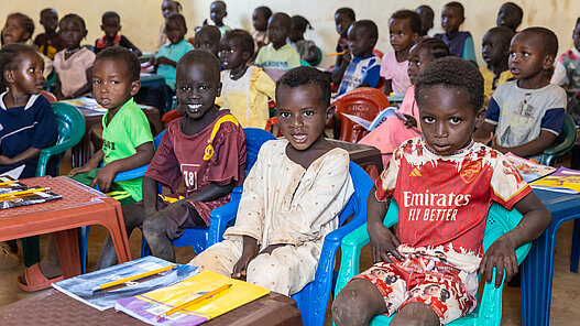 Guppe von Kindern sitzt in einem Klassenzimmer. Im Vordergrund sind vier Jungs zu sehnen die vor ihren Schulheften sitzen.