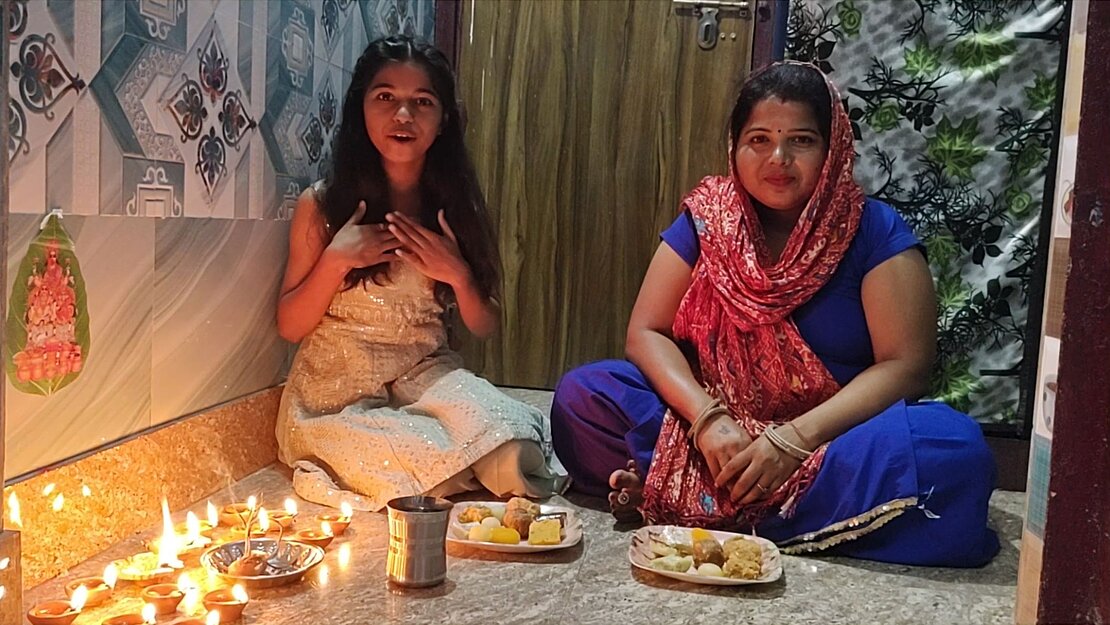 Ein Mädchen und eine Frau sitzen in einem Raum auf dem Fußboden, vor ihnen stehen zwei Teller mit Gebäck und im Raum sind viele Kerzen angezündet