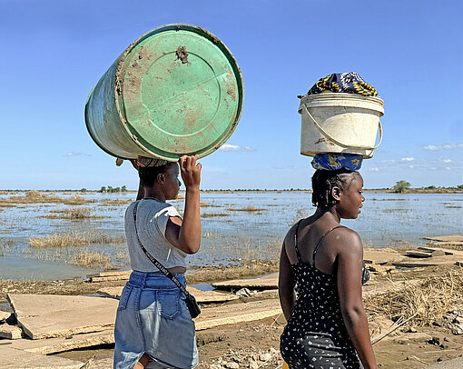 Zwei Frauen tragen Güter auf einen Kopf und laufen durch ein überschwemmtes Feld