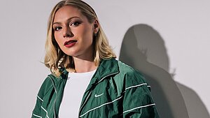 Halbporträt einer Frau mit schulterlangem blondem Haar, die eine grüne Sportjacke über einem weißen Oberteil trägt. Sie steht vor einer hellen Wand.