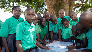Schülergruppe in grünen T-Shirts diskutiert im Freien im Rahmen eines Schulgesundheitsclubs in Uganda über Menstruationshygiene, Jungen und Mädchen arbeiten gemeinsam an einer Zeichnung mit Empfehlungen zu Hygienepraktiken.