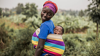 Eine Mutter trägt ihr Baby auf dem Rücken. Das Baby gähnt und die Mutter schaut liebevoll nach hinten. 