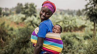 Eine Mutter trägt ihr Baby auf dem Rücken. Das Baby gähnt und die Mutter schaut liebevoll nach hinten. 