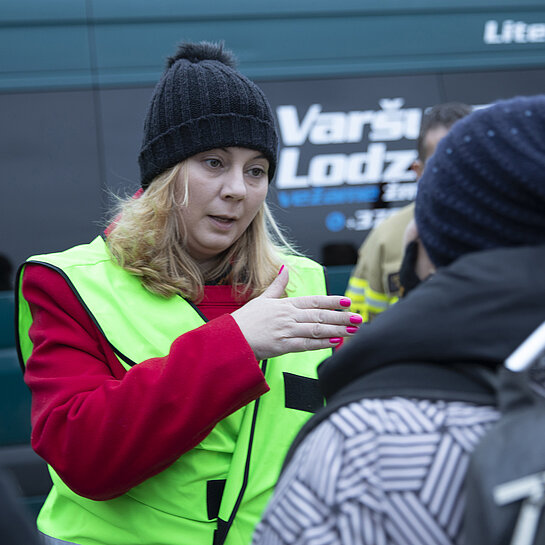 Monica steht in gelber Warnweste vor einem Bus und spricht mit Menschen.