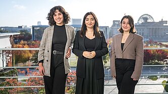 Zwei junge Frauen stehen an der Seite der Integrationsstaatsministerin auf der Terasse der Kanzleramts.