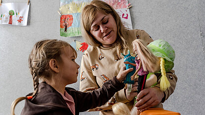 Bild: Ein kleines Kind spielt mit einer Puppe, die von ihrer Betreuerin gehalten wird
