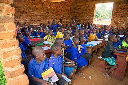 In einem Klassenraum sitzen über 40 Kinder eng nebeneinander auf Holzbänken. Sie tragen blaue Schuluniformen und schauen nach vorne in Richtung Tafel. 