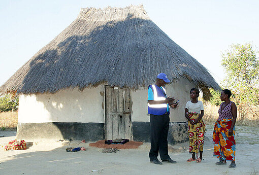 Plan-Mitarbeiter mit zwei Frauen vor einer Lehmhütte mit Strohdach