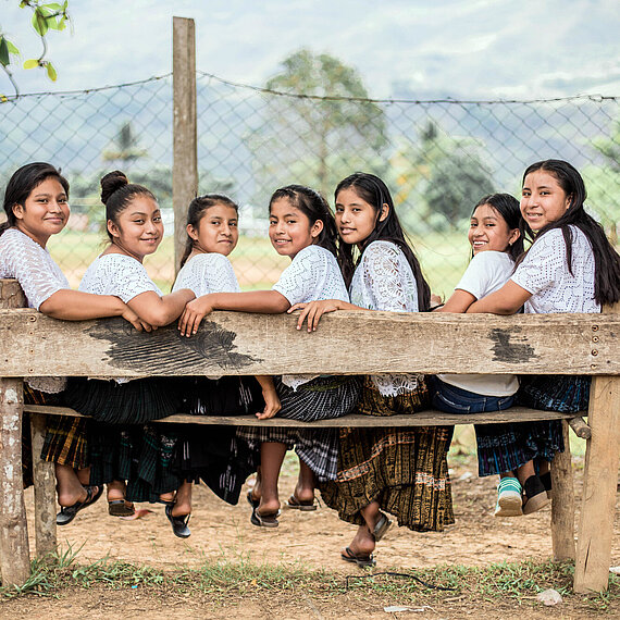 Sieben Mädchen einer von Plan gegründeten Leadership School in Guatemala sitzen auf einer Bank und lächeln in die Kamera Gruppe von sieben Mädchen in traditioneller Kleidung, die auf einer Bank im Freien sitzen und in die Kamera lächeln, mit einem Zaun und einer grünen Landschaft im Hintergrund, in Guatemala.