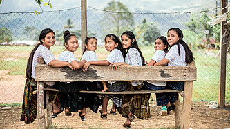 Gruppe von sieben Mädchen in traditioneller Kleidung, die auf einer Bank im Freien sitzen und in die Kamera lächeln, mit einem Zaun und einer grünen Landschaft im Hintergrund, in Guatemala.