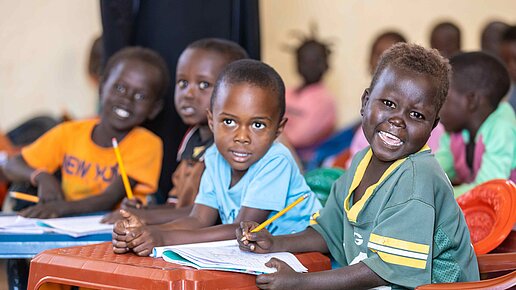 Zwei Jungen sitzen im Vordergrund in einem Klassenzimmer. Der eine hat einen Stift in der Hand und guckt sprechend in die Kamera. Beide haben Hefte vor sich liegend.