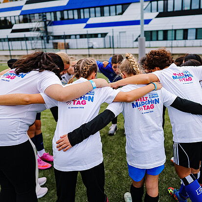 Eine Gruppe von Mädchen in weißen T-Shirts mit der Aufschrift "Weltmädchentag" steht im Kreis und hält sich in den Armen. Sie stecken die Köpfe zusammen, wie eine Fußballmannschaft, die sich gegenseitig anfeuert.