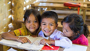 Drei Schulkinder aus Laos in einem Klassenzimmer zeigen auf ein Buch und lächeln.