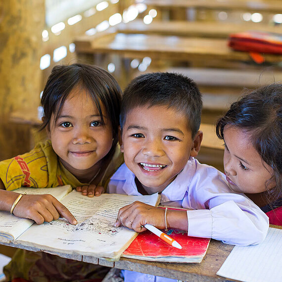 Drei Schulkinder aus Laos in einem Klassenzimmer zeigen auf ein Buch und lächeln.