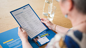 Eine ältere Person hält eine Checkliste zur Nachlassplanung von Plan International in den Händen und sitzt an einem Holz-Tisch. Neben dem blauen Ordner liegen ein Glas Wasser und eine Wasserflasche. Das Dokument enthält verschiedene Kategorien zum Thema Vorsorge.