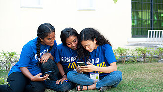 Drei junge Frauen sitzen auf einer Wiese und schauen auf ein Smartphone