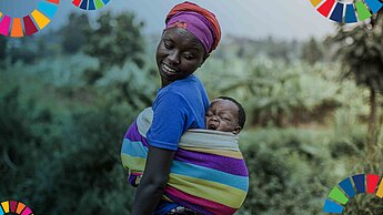Eine Frau trägt ein kleines Kind mit einem Tragetuch auf ihrem Rücken und lächelt es über die Schulter schauend an.
