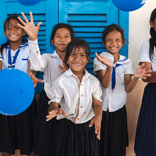 Fröhliche Schulkinder in Schuluniformen stehen vor einer blauen Wand und spielen lachend mit blauen Luftballons.