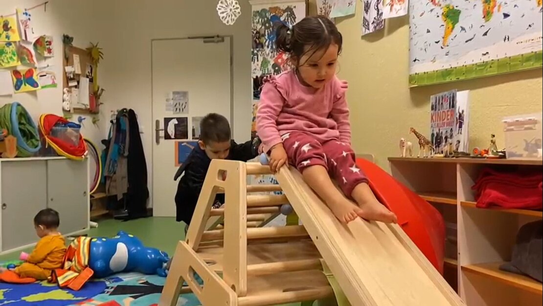 Kinder spielen in einem bunten Spielzimmer, ein kleines Mädchen rutsch von einer kleinen Holzrutsche