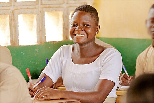 Damipi lernt im Unterricht an ihrer Schule im Norden Togos. Ein Mädchen sitzt an einem Tisch, hat einen Stift in der Hand und lächelt breit.