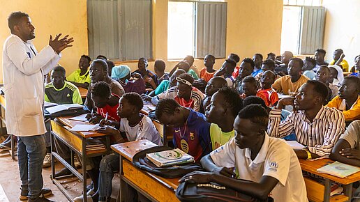 Ein Lehrer steht gestikulierend (etwas erklärend) vor einem voll besetzten Klassenzimmer mit Jugendlichen.