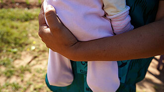 Eine Nahaufnahme von einem gebräunten Arm, der ein Baby in einem rosa Strampler hält. 