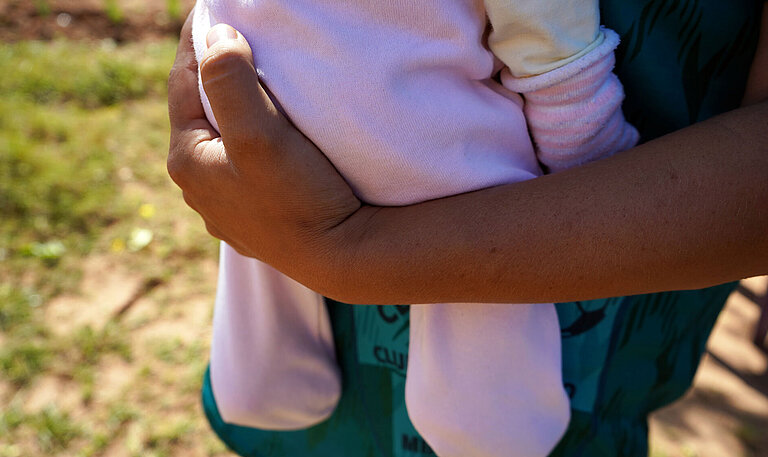 Eine Nahaufnahme von einem gebräunten Arm, der ein Baby in einem rosa Strampler hält. 