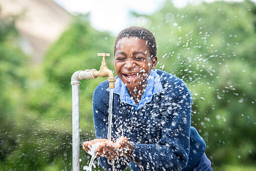 Ein Mädchen in Schuluniform hält seine Hände unter einen laufenden Wasserhahn. Das Wasser spritzt in alle Richtungen und das Mädchen lacht.