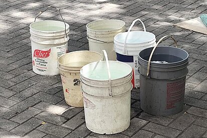 Mehrere Eimer stehen auf einer gepflasterten Fläche, im Hintergrund leben Menschen nach dem Erdbeben in Zelten auf der Straße in Caracas.