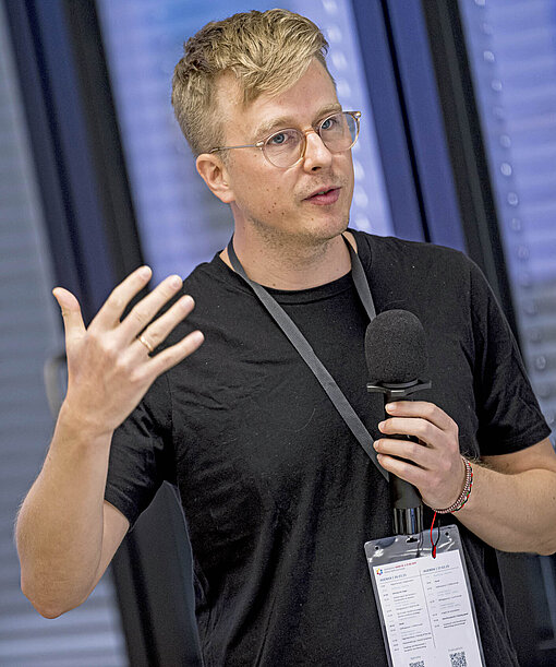 Ein Mann im schwarzen T-Shirt steht in einem Konferenzraum und spricht