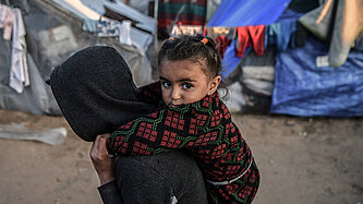Ein kleines Mädchen sitzt auf dem Rücken eines Erwachsenen in einem Geflüchtetenlager, mit einem Zelt und Wäsche im Hintergrund.