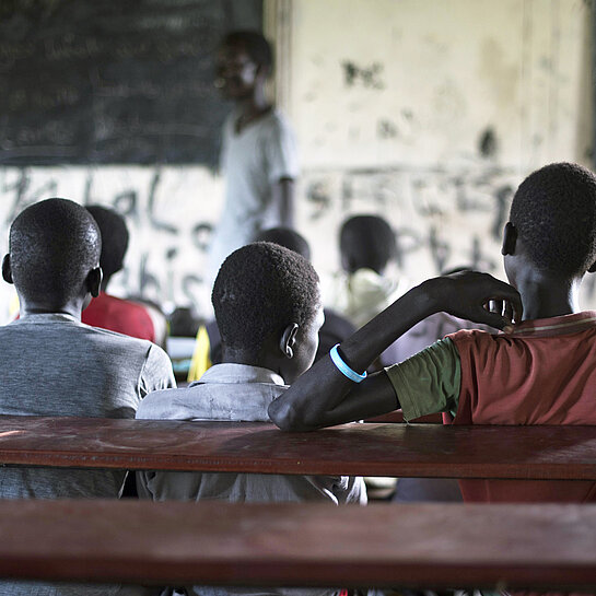 Auf einer hölzernen Schulbank sitzen in einem dunklen Klassenzimmer drei Jungen in T-Shirts nebeneinander und blicken zur Tafel und dem Lehrer